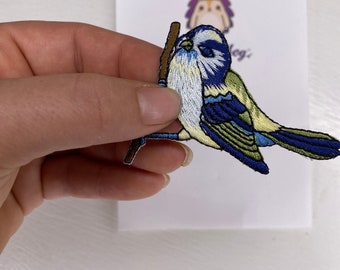 Puede incluir: Un parche bordado para planchar de un pájaro azul que sostiene un palo en su pico. El pájaro es azul, blanco y verde con un palo marrón.