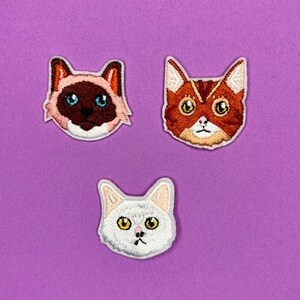 Puede incluir: Tres parches bordados con forma de cara de gato sobre un fondo morado. Los parches son de un gato siamés, un gato atigrado y un gato blanco.