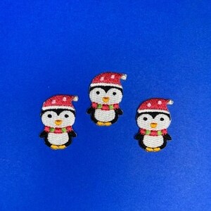 Puede incluir: Tres parches bordados de pingüinos con gorros rojos de Papá Noel y bufandas blancas. Los pingüinos están sobre un fondo azul.