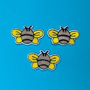 May include: Three embroidered bee patches with yellow wings and a gray body. The bees are on a blue background.