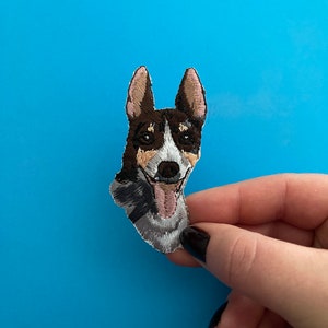 May include: Embroidered patch of a brown and white dog with a pink tongue sticking out. The dog is looking directly at the camera and has a happy expression.