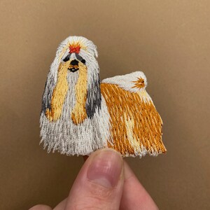 May include: Embroidered patch of a Shih Tzu dog with white, brown, and black fur. The dog is looking directly at the viewer.