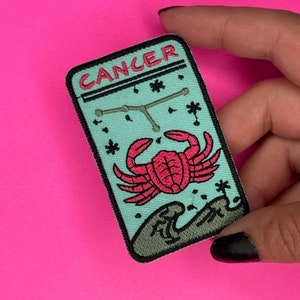 Op de afbeelding: Een turquoise blauwe geborduurde patch met het woord "Kanker" in zwarte en roze letters. De patch toont een roze krab met zwarte contouren en een sterrenbeeld. De krab zit op een golf.