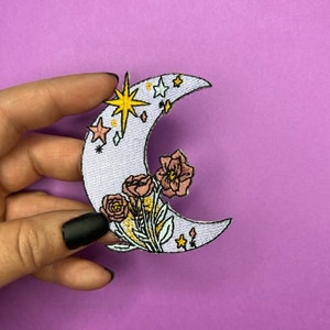 May include: A white crescent moon embroidered patch with pink flowers, green leaves, and yellow stars. The moon has a light purple outline.