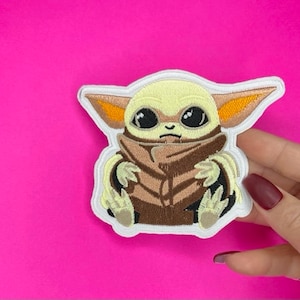 Puede incluir: Un parche bordado con un personaje de la franquicia Star Wars, conocido como "The Child" o "Baby Yoda". El personaje lleva una túnica marrón y tiene grandes ojos oscuros.