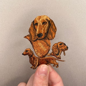 May include: An embroidered patch featuring three brown dachshund dogs. The dogs are in different poses, with one standing, one sitting, and one lying down. The patch is made of brown thread on a white background.