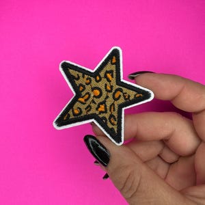 May include: A brown and orange star-shaped patch with a leopard print design. The patch has a white outline and is embroidered.