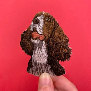 May include: Embroidered patch of a brown and white dog with floppy ears, looking up. The dog has a pink tongue sticking out.