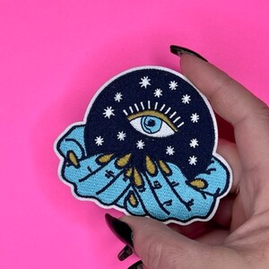 Puede incluir: Un parche bordado azul y blanco con un ojo celestial rodeado de estrellas, sostenido por dos manos con u&ntilde;as doradas.