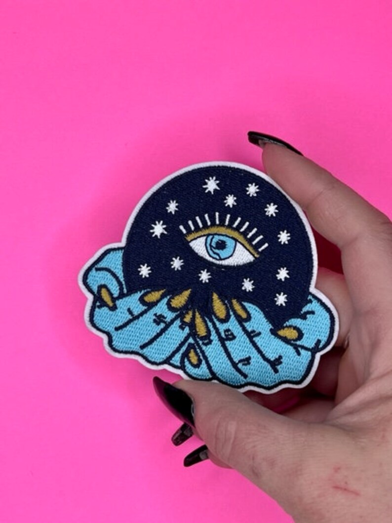Puede incluir: Un parche bordado azul y blanco con un ojo celestial rodeado de estrellas, sostenido por dos manos con u&ntilde;as doradas.