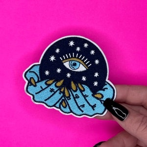 Peut inclure: Un patch brodé bleu et noir avec un œil stylisé entouré d'étoiles et une main tenant une sphère. Le patch est sur un fond rose.