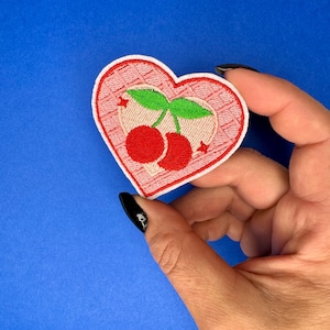 May include: A pink heart-shaped embroidered patch with two red cherries and green leaves. The cherries have white stars on them.