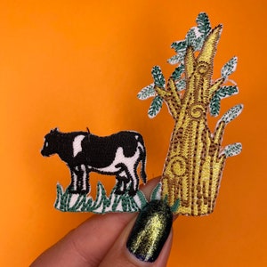 May include: Black and white cow embroidered patch with green grass and a brown and yellow tree with green leaves.
