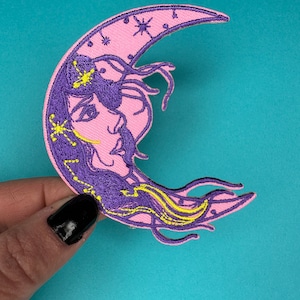 Può includere: Un cerchio di luna rosa a forma di mezzaluna con un viso di donna viola e stelle gialle ricamate.