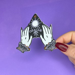 May include: Black and white embroidered patch of two hands holding a black and white ouija board with the word "ORACLE" written on it.