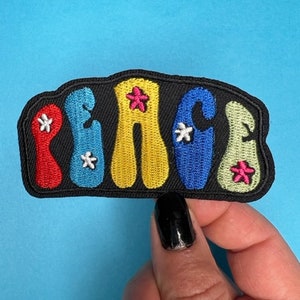 May include: A colorful embroidered patch with the word "PEACE" in a retro font. The letters are outlined in black and filled with red, blue, yellow, and green. There are small white and pink stars scattered throughout the patch.