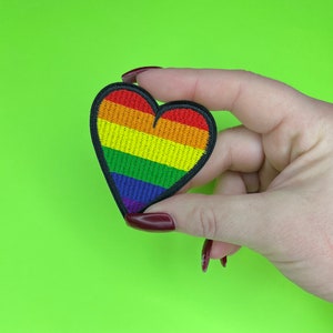 May include: A rainbow heart-shaped patch with red, orange, yellow, green, blue, indigo, and violet stripes. The patch is held in a hand.