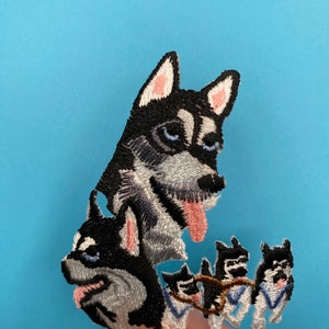 Pode incluir: Etiqueta bordada com um cão husky preto e branco com a língua rosa de fora. O husky é cercado por quatro cães husky menores, todos puxando um trenó.