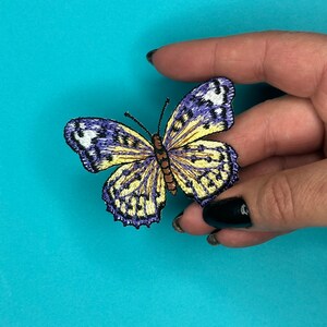 May include: A yellow and purple embroidered butterfly patch with brown accents. The butterfly is in flight with its wings spread open.