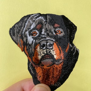 May include: Embroidered patch of a Rottweiler dog's face. The dog has black and brown fur with a white patch on its forehead. The patch is made of black fabric with detailed embroidery.