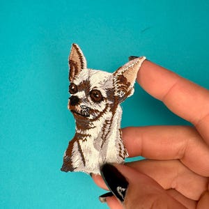 May include: A white and brown embroidered chihuahua patch. The patch is detailed with brown ears, a brown nose, and brown eyes.