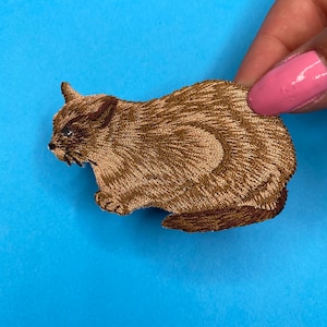 May include: A brown embroidered iron-on patch of a cat with a grumpy expression.