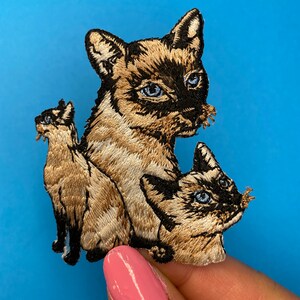 May include: Three embroidered iron-on patches of Siamese cats in various poses. The patches are beige, brown, and black.