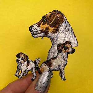 May include: Embroidered patch featuring three Jack Russell Terriers. The largest dog is brown and white with its head turned to the side. The two smaller dogs are white with brown markings. The patch is on a yellow background.