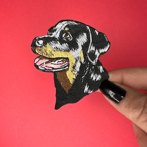 May include: Embroidered patch of a Rottweiler dog's head, with black, brown, and white fur. The dog is looking to the left, with its mouth open and tongue sticking out.