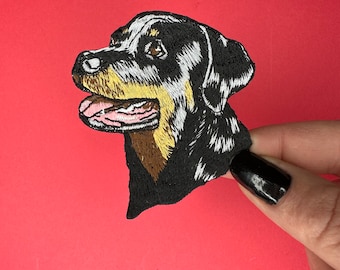 May include: Embroidered patch of a Rottweiler dog's head, with black, brown, and white fur. The dog is looking to the left, with its mouth open and tongue sticking out.