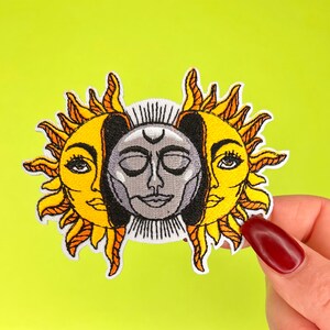 May include: Embroidered patch featuring a sun and moon design. The sun and moon are depicted as faces, with the sun having a yellow face and the moon having a gray face. The sun and moon are intertwined, with the sun's rays extending around the moon.