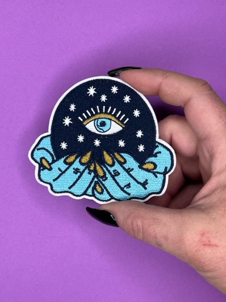 Puede incluir: Un parche bordado azul y negro con un ojo blanco rodeado de estrellas, sostenido en dos manos con anillos dorados. Las manos sostienen una esfera azul oscuro con estrellas blancas.