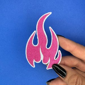 Könnte beinhalten: Ein pinkfarbener, gestickter Flammenaufnäher mit weißem Rand. Das Flammendesign hat eine leuchtend pinke Farbe. Der Aufnäher wird vor einem blauen Hintergrund gehalten.