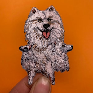 Puede incluir: Parche bordado de un perro blanco y esponjoso con la lengua rosa que sobresale, sosteniendo dos perros blancos y esponjosos más pequeños en sus patas.