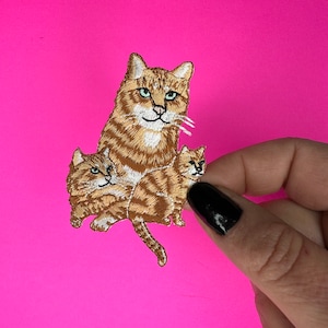 May include: Embroidered patch featuring three orange tabby cats. The mother cat is sitting upright with her two kittens nestled beside her.