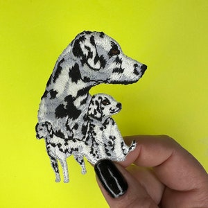 May include: Embroidered patch of a Dalmatian dog with a smaller Dalmatian puppy nestled against it. The patch is white with black spots and has a detailed design.