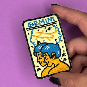 Puede incluir: Un parche bordado amarillo y blanco con la palabra "GEMINI" en letras negras. El parche presenta una ilustración de dibujos animados de dos personas con cabello azul y piel amarilla, con una constelación de estrellas sobre ellos.