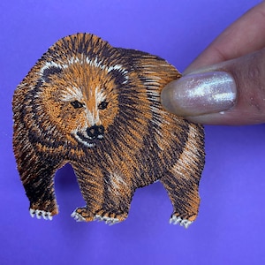 Peut inclure: Patch brodé en forme d'ours brun avec des détails noirs et blancs. L'ours est debout sur ses quatre pattes et a une apparence réaliste.