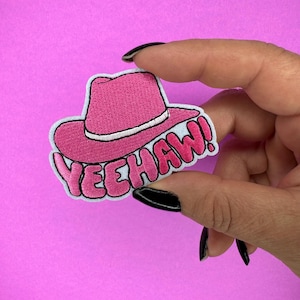 Puede incluir: Un parche bordado rosa con un diseño de sombrero de vaquero y la palabra "YEEHAW!" en letras de burbujas. El parche tiene un borde blanco, sobre un fondo rosa.
