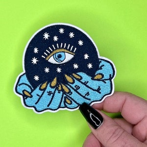 Puede incluir: Un parche bordado azul y negro con un ojo grande rodeado de estrellas y una mano sosteniendo una esfera. El ojo es azul con una pupila blanca y un iris negro.