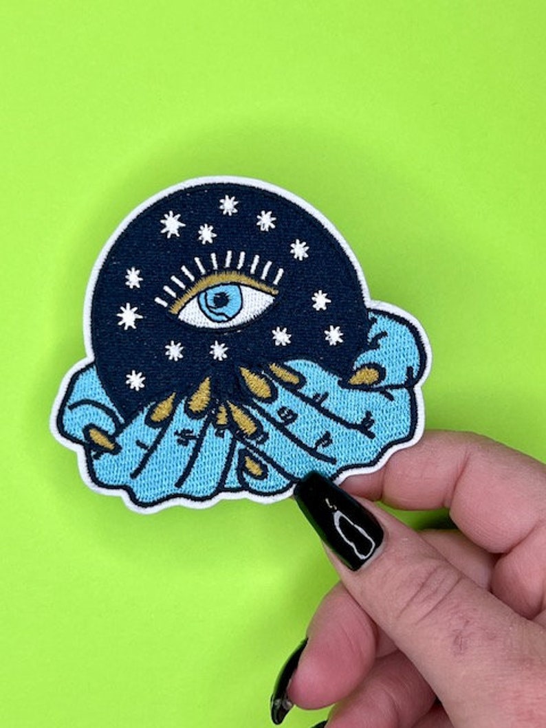 Puede incluir: Un parche bordado azul y negro con un ojo grande rodeado de estrellas y una mano sosteniendo una esfera. El ojo es azul con una pupila blanca y un iris negro.