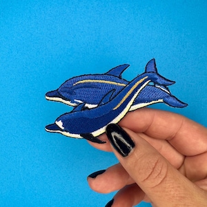 May include: Two blue embroidered dolphin patches with yellow accents. The patches are held in a hand with black nail polish.