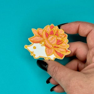 以下が含まれることがあります： 白い刺繍のアイロン接着パッチ。頭の上にピンクとオレンジの花をつけたハリネズミの模様です。
