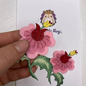 Op de afbeelding: Twee roze viltbloem-patches met rode centra en gele knoppen. De bloemen zijn geborduurd met groene bladeren en stelen.