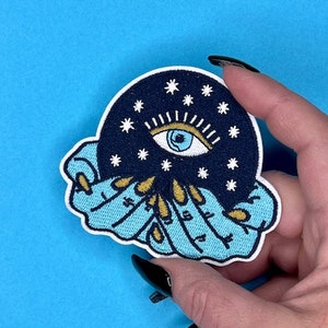 Puede incluir: Un parche bordado azul y negro con un ojo estilizado rodeado de estrellas y un anillo de manos con detalles dorados.