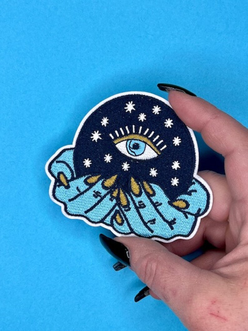 Puede incluir: Un parche bordado azul y negro con un ojo estilizado rodeado de estrellas y un anillo de manos con detalles dorados.