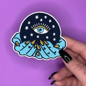 Puede incluir: Un parche bordado azul y negro con un gran ojo rodeado de estrellas, sostenido en dos manos con detalles dorados.