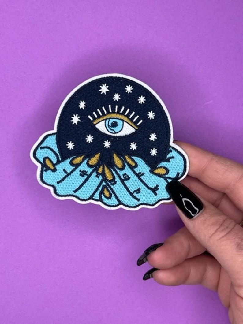 Puede incluir: Un parche bordado azul y negro con un gran ojo rodeado de estrellas, sostenido en dos manos con detalles dorados.