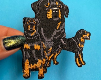 May include: Embroidered patch featuring three Rottweilers in different sizes. The largest Rottweiler is black with brown markings and is standing with its head tilted slightly to the side. The medium-sized Rottweiler is also black with brown markings and is standing behind the largest Rottweiler. The smallest Rottweiler is black with brown markings and is standing in front of the other two Rottweilers.