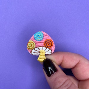 以下が含まれることがあります： ピンクのキノコにカラフルな渦巻きが刺繍されています。キノコは小さな装飾用パッチです。